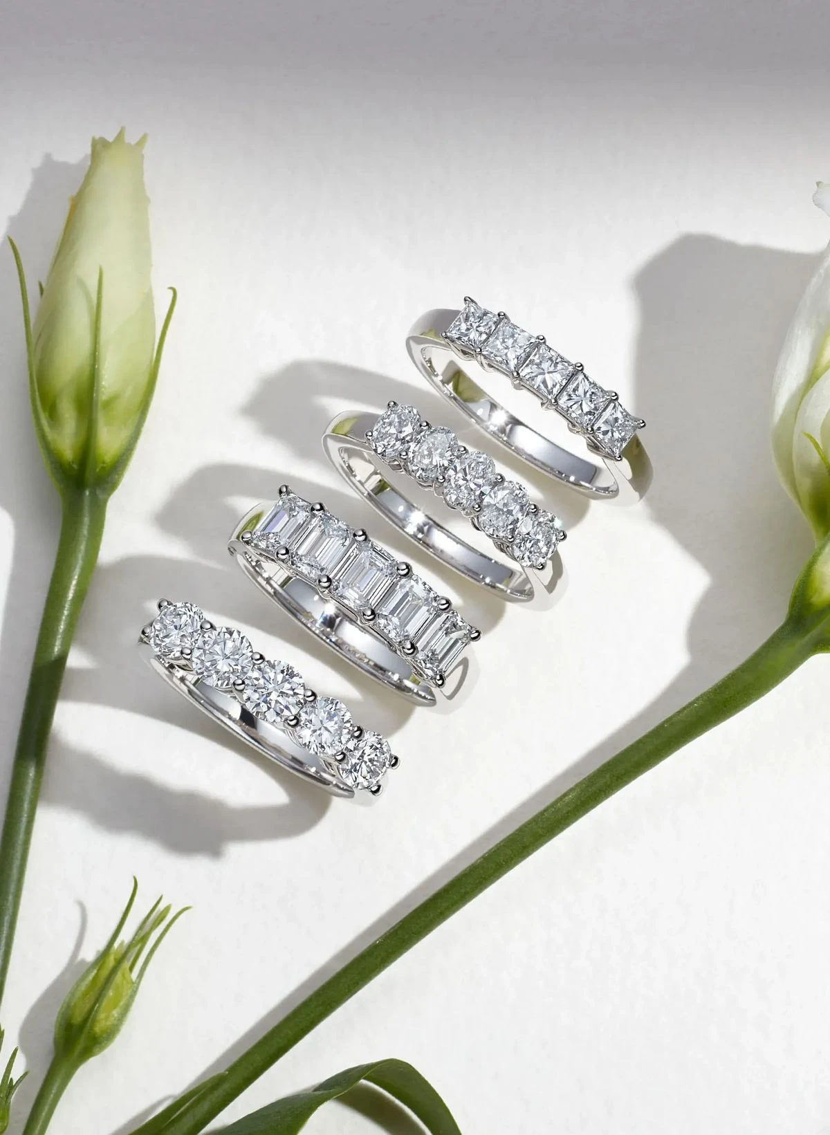 Four diamond bands with different stone shapes arranged beside green flower stems
