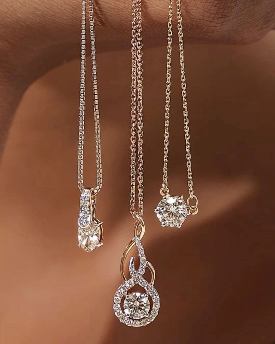 Three diamond pendant necklaces hanging from fine chains against a hand